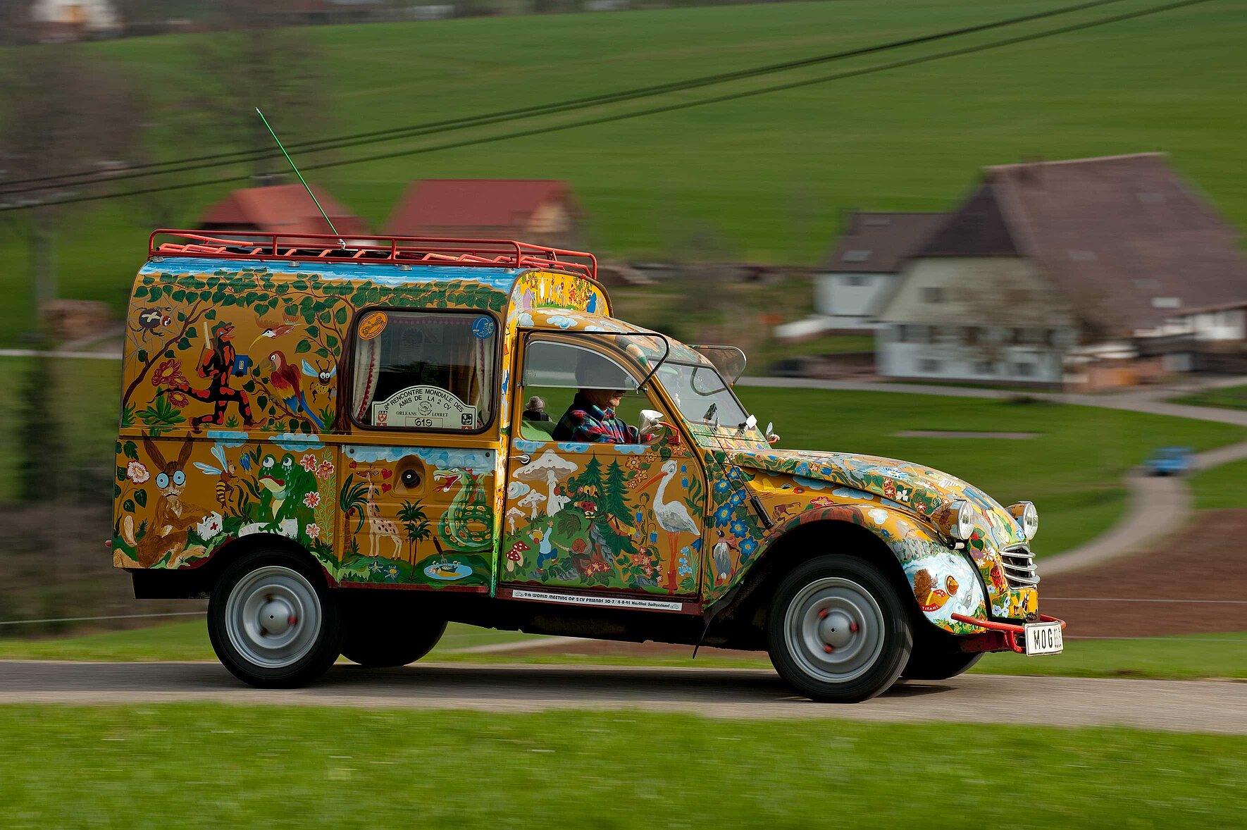 Ein über Land fahrender Oldtimer, der mit verschiedensten Tieren und Fabelwesen bemalt ist.
