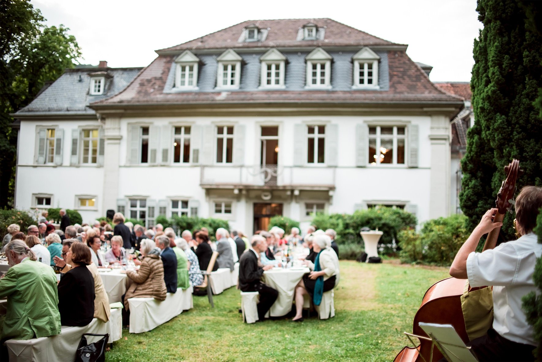Festlich gekleidete Menschen der Stiftung der Freunde Heidelberg sitzen an gedeckten Tischen in einem Garten und essen zusammen. Ein Mann spielt Cello. 
