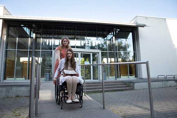 Eine Frau im Rollstuhl wird von einer anderen Frau eine Rampe vor einem Gebäude hinuntergeschoben.