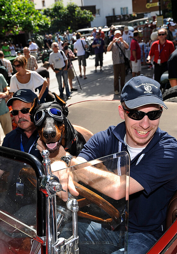 Der Sohn des Autosammlers, Hannes Steim, mit seinem Hund und seinem Vater in einem Oldtimer unterwegs. Viele Passant:innen schauen zu.